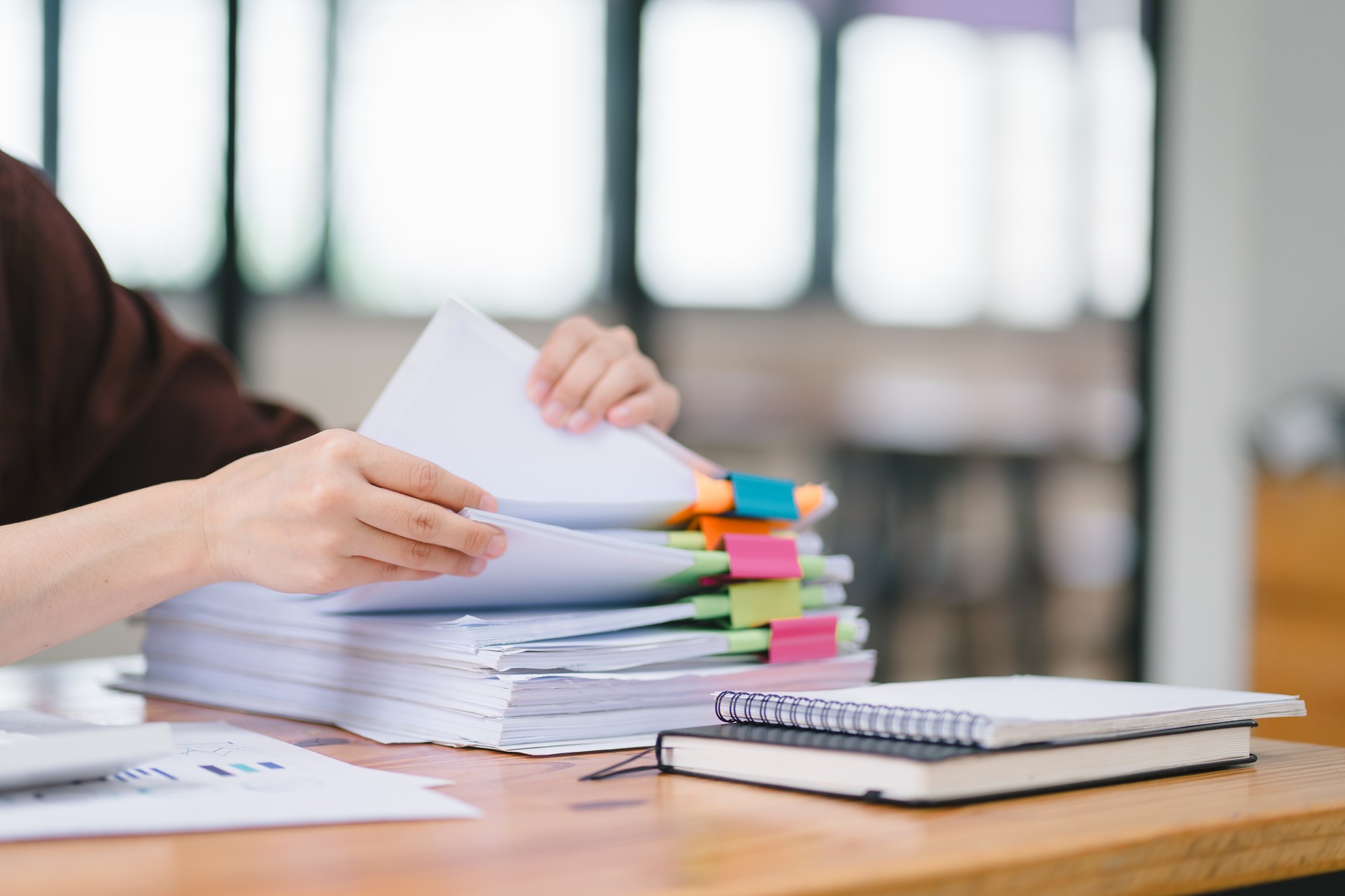 The businesswoman's hands are busy working amidst stacks of paper files, searching and checking for unfinished documents among the folders and papers on her cluttered office desk.